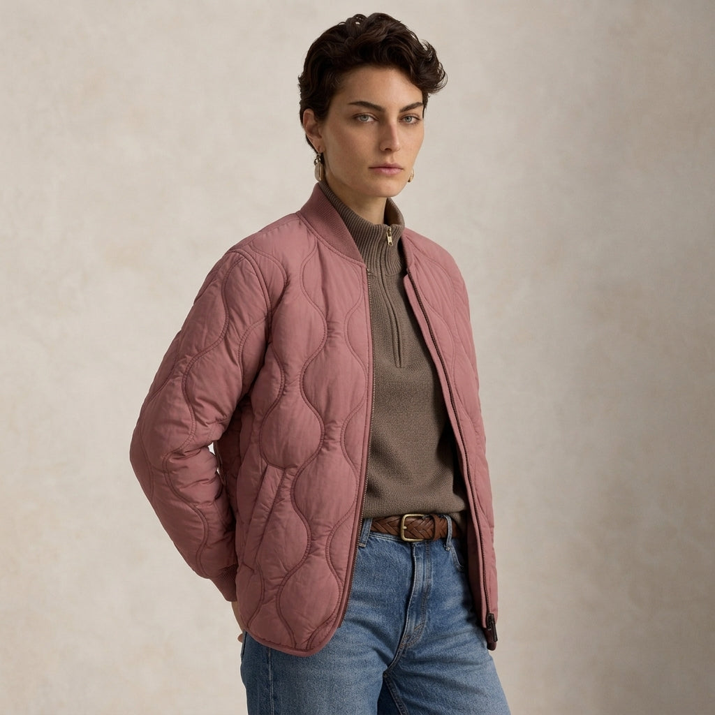 A woman wearing a Quilted Pink Jacket over a taupe turtleneck sweater and blue jeans, standing against a textured beige studio backdrop.