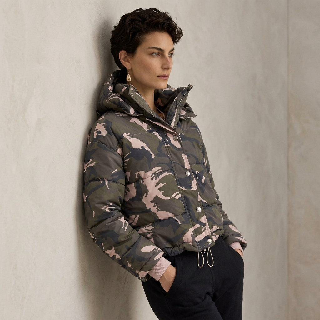 A woman wearing a Quilted Camo Jacket with pink and gray tones, leaning against a textured light gray wall.