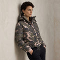 A woman wearing a Quilted Camo Jacket with pink and gray tones, leaning against a textured light gray wall.