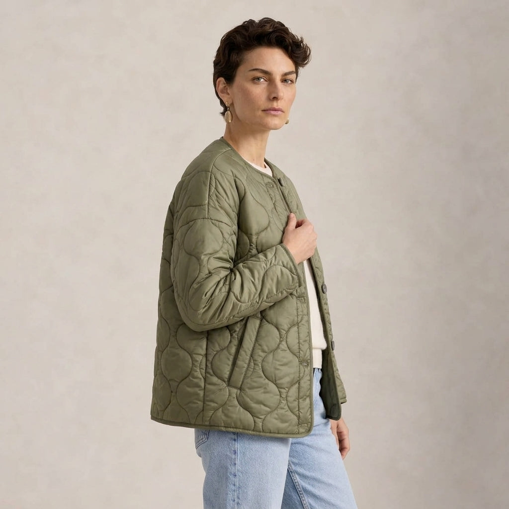 A woman wearing an olive green quilted jacket, styled with a white t-shirt and light blue jeans, standing against a neutral textured beige background.