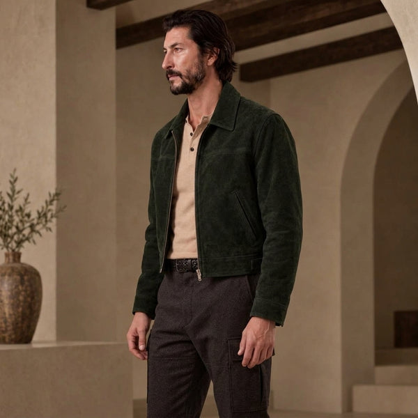 A man wearing a dark green Suede Short Jacket over a beige polo shirt and dark gray trousers, standing in a warm-toned architectural interior with arches and a vase.