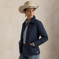 A woman wearing a navy blue diamond-Western Quilted Jacket, a light blue button-up shirt, blue jeans, and a beige cowboy hat, posed against a textured beige studio background.