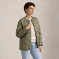 A woman wearing an olive green Satin Quilted Jacket over a cream sweater and light blue jeans, standing against a textured beige studio background.