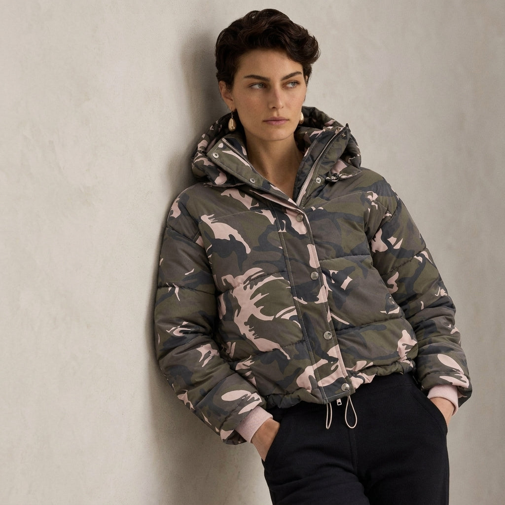A woman wearing a camouflage hooded puffer jacket leans against a textured light gray wall. The jacket features a green, gray, and beige camo pattern with a drawstring hood and front zipper.