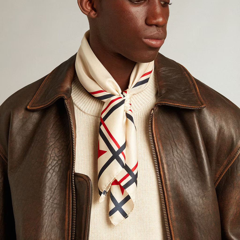 A close-up of a model wearing a cream silk scarf with a red and navy blue plaid pattern, styled under a brown leather jacket and cream knit sweater.