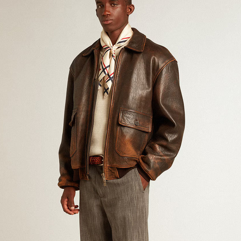 A male model wearing a distressed brown leather aviator jacket over a cream sweater and a patterned neck scarf, paired with gray trousers, against a plain light gray studio background.