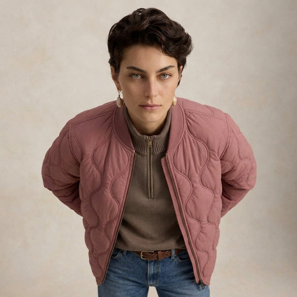 A model wearing a dusty rose pink quilted bomber jacket over a brown ribbed half-zip sweater and blue jeans, set against a neutral beige textured background.