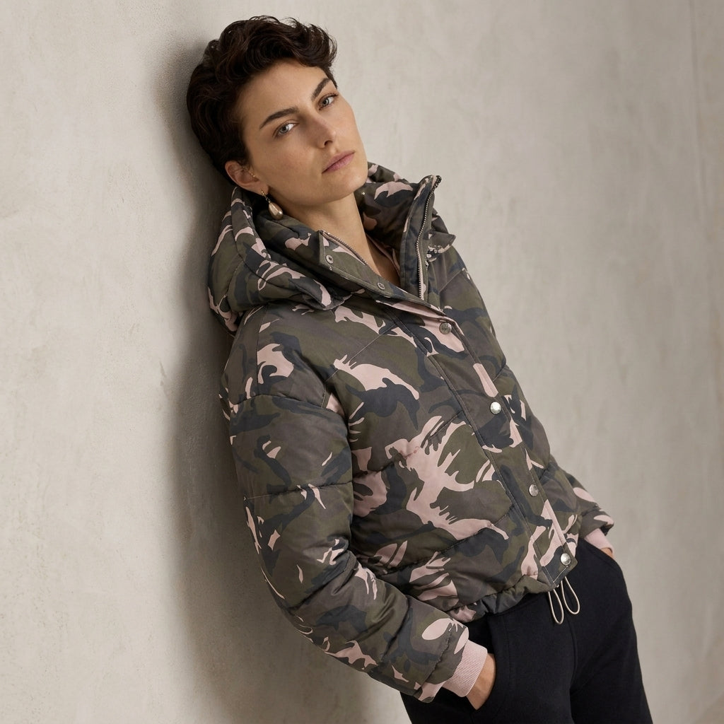 A model wearing a camouflage puffer jacket with a hood, leaning against a textured light gray wall.