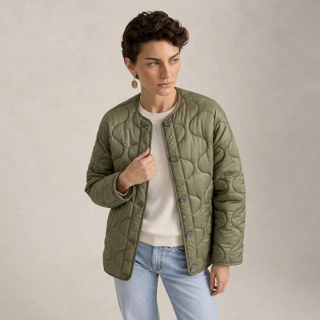 A woman wearing an olive green quilted jacket over a cream sweater and light blue jeans, standing against a neutral beige studio background.