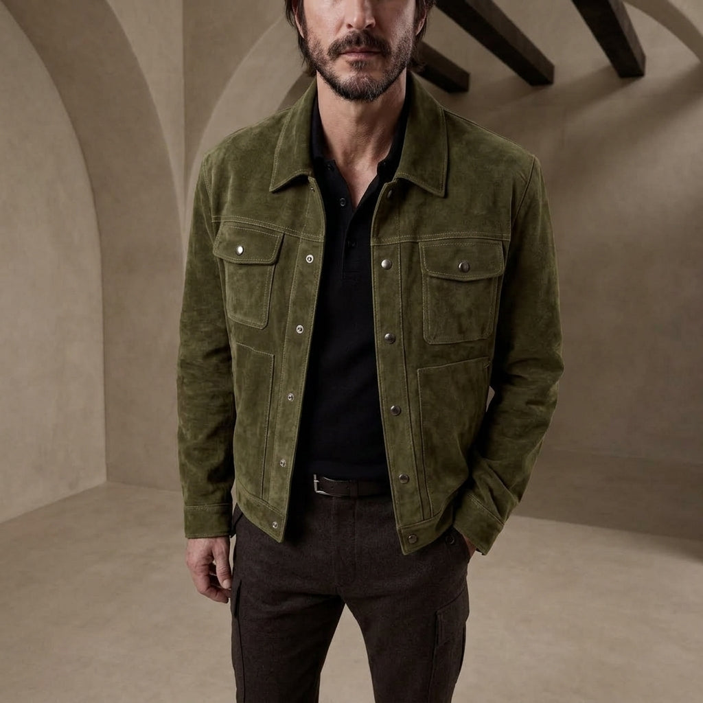 A man wearing an olive green suede jacket over a black polo shirt and dark brown trousers, standing in a warm-toned stone room with arched walls and wooden ceiling beams.