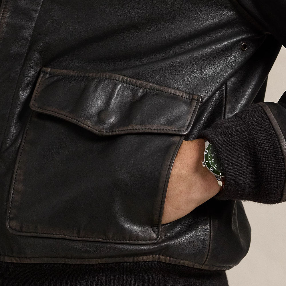Close-up of a black leather jacket with a large flap pocket and a person's hand in the pocket, wearing a dark ribbed knit sleeve and a wristwatch.