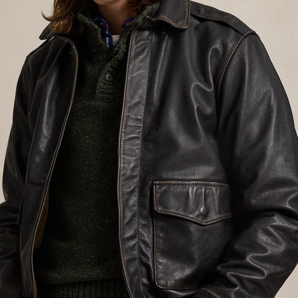 A close-up of a person wearing a dark brown leather jacket over a dark green knit sweater, against a plain light beige background.