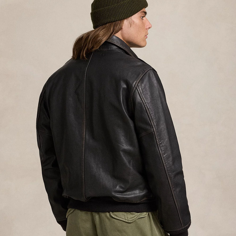 A man wearing a black leather jacket, olive green pants, and an olive green beanie, photographed from the back against a plain beige studio background.