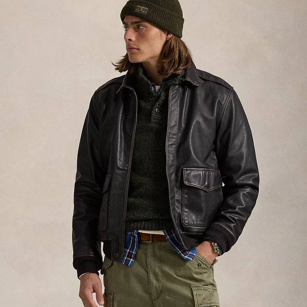 A man wearing a black leather aviator-style jacket over a dark knit sweater, with an olive green beanie, plaid shirt, and cargo pants, standing against a plain beige studio background.