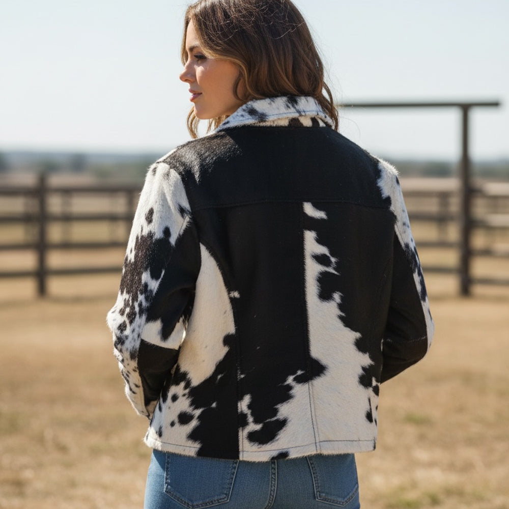A woman wearing a brown and white cowhide jacket, shown from the back in an outdoor ranch setting with a fence in the background.