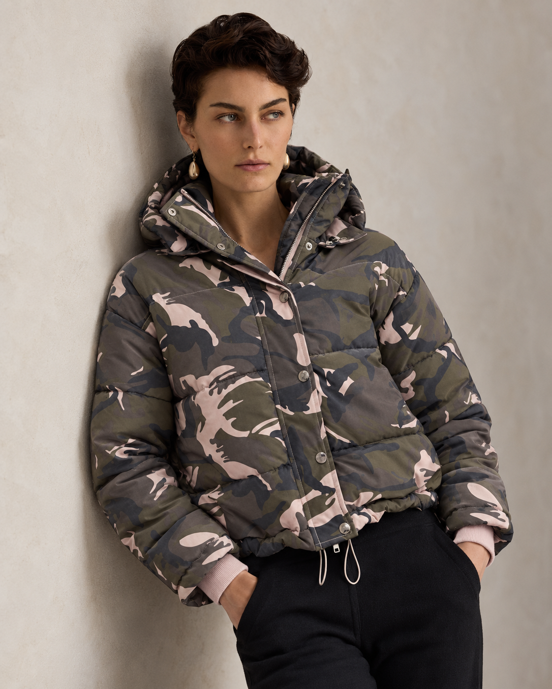 A woman wearing a hooded puffer jacket with a green, gray, and beige camouflage pattern, paired with black pants, leaning against a textured light gray wall.