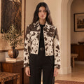 A woman stands in a warmly lit interior wearing a cropped cowhide jacket with a brown and cream cow-print pattern over a black top and black trousers.