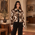 A woman stands indoors wearing a cropped fringe jacket with a brown and cream cow-print pattern over a black outfit. The room has warm neutral walls, framed artwork, a wooden sideboard, and a rug.