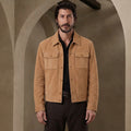 A man wearing a tan suede jacket over a black shirt and dark trousers, standing in front of a warm-toned arched wall.