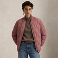 A model wearing a dusty rose quilted bomber jacket over a brown ribbed zip-up sweater and blue jeans, standing against a textured beige wall.