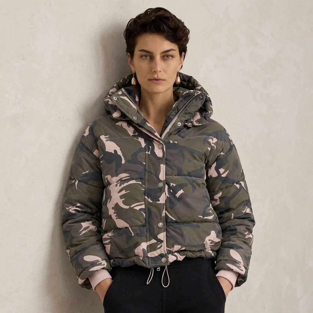A model wearing a cropped camouflage puffer jacket with a hood, standing against a textured light gray wall.