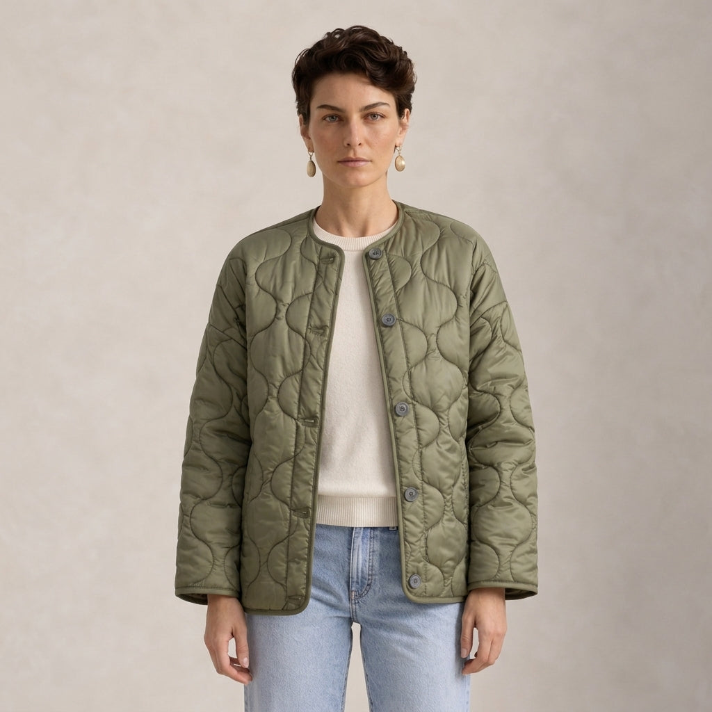 A woman modeling a quilted olive green jacket over a cream sweater and light blue jeans, standing against a neutral textured beige background.