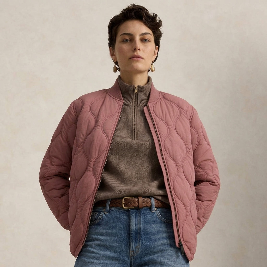 A woman wearing a dusty rose quilted bomber jacket over a taupe quarter-zip sweater and blue jeans, standing against a textured beige background.