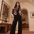 A woman stands in a warm, elegant interior wearing a cropped Cowhide Trucker Jacket over a black top and black wide-leg trousers.
