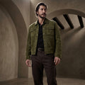 A man wearing an olive Green Suede Trucker Jacket over a black shirt and dark brown pants, standing in a rustic stone room with arches.