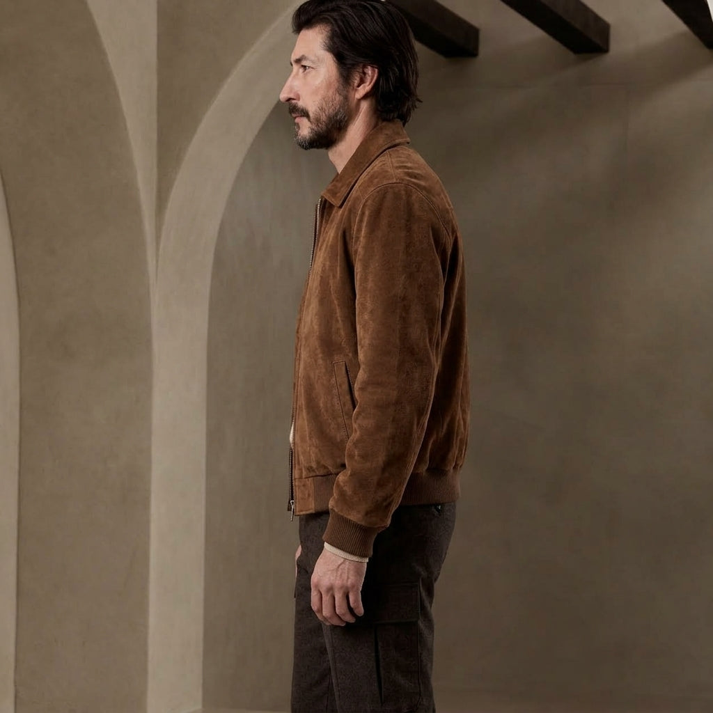 A man in side profile wearing a brown suede jacket and dark trousers, standing in a warm-toned interior with arched walls.