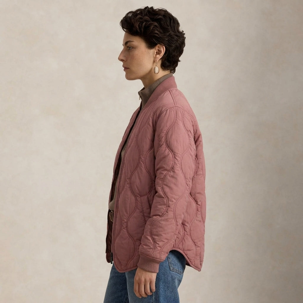 A woman in profile wearing a dusty rose quilted jacket over a brown collared shirt and blue jeans, standing against a textured beige studio background.