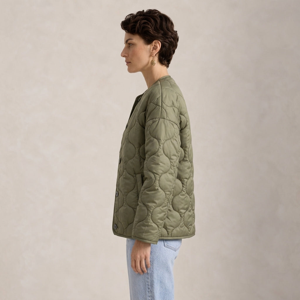 A woman in profile wearing an olive green quilted jacket and light blue jeans, standing against a textured beige studio background.
