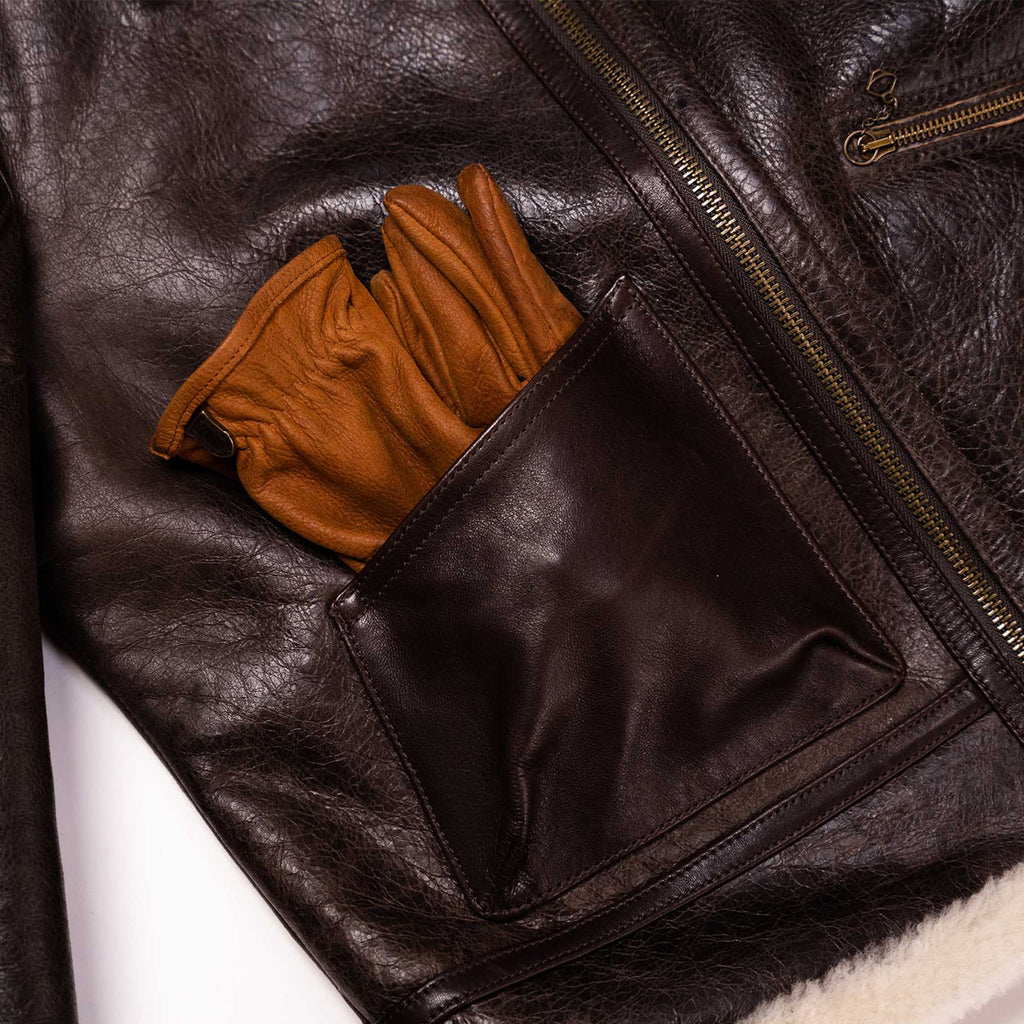 Close-up of a dark brown leather jacket with a side pocket holding a pair of tan leather gloves, showing the texture and details of the jacket's construction.