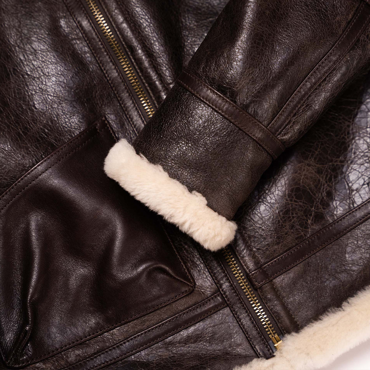 Close-up of a dark brown leather jacket with a cream-colored shearling lining visible at the cuff and hem, featuring gold-toned zippers.