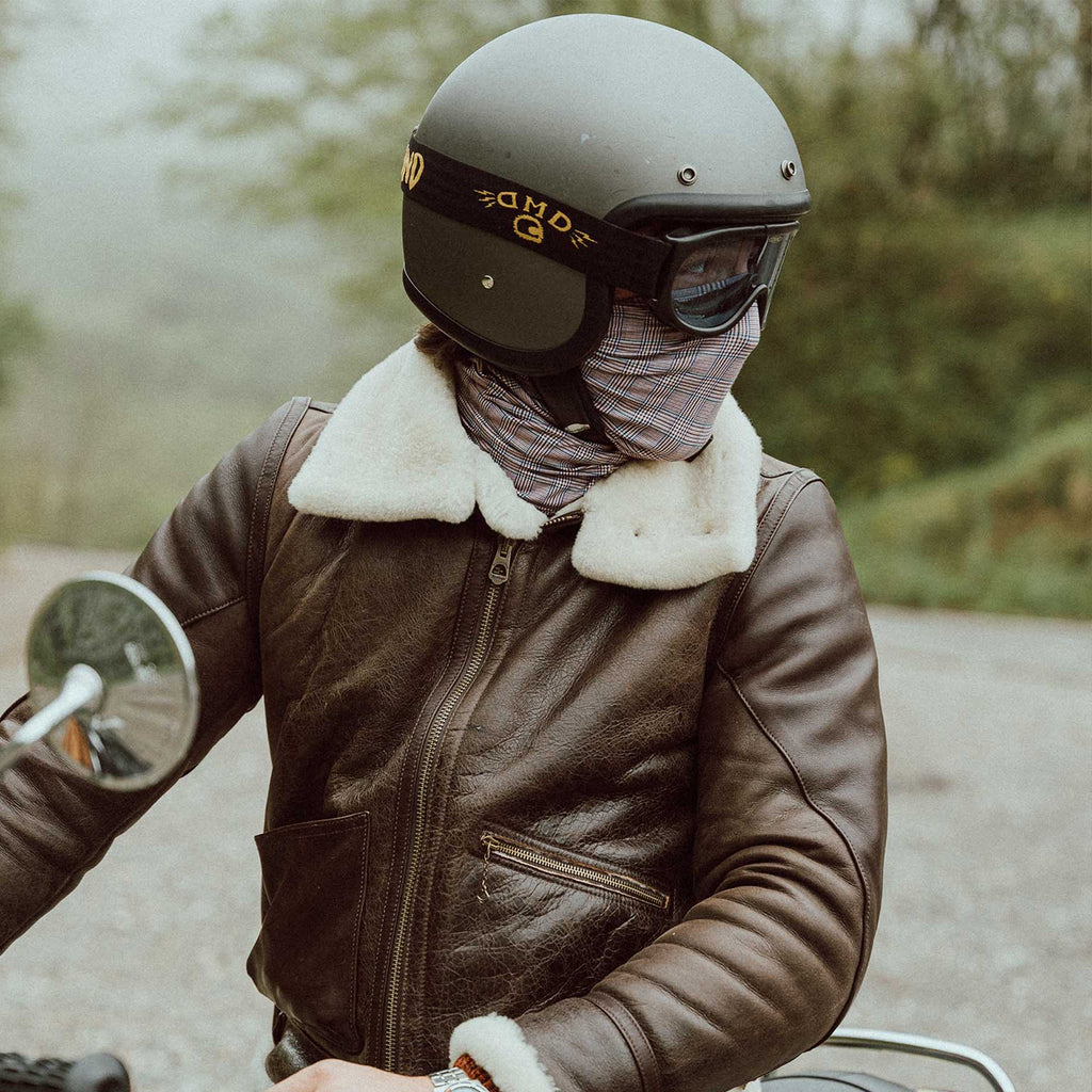 A person wearing a dark brown leather aviator-style jacket with a white shearling-style collar, a gray helmet, and a face covering, riding a motorcycle outdoors on a misty road.