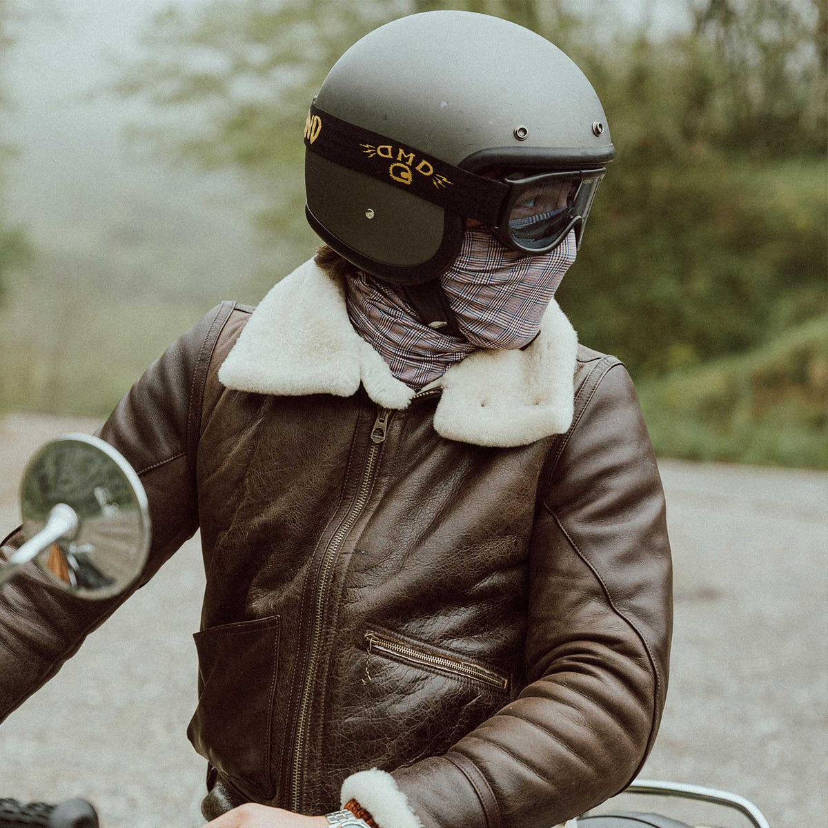 A person wearing a dark brown leather aviator-style jacket with a white shearling-style collar, a gray helmet, and a face covering, riding a motorcycle outdoors on a misty road.
