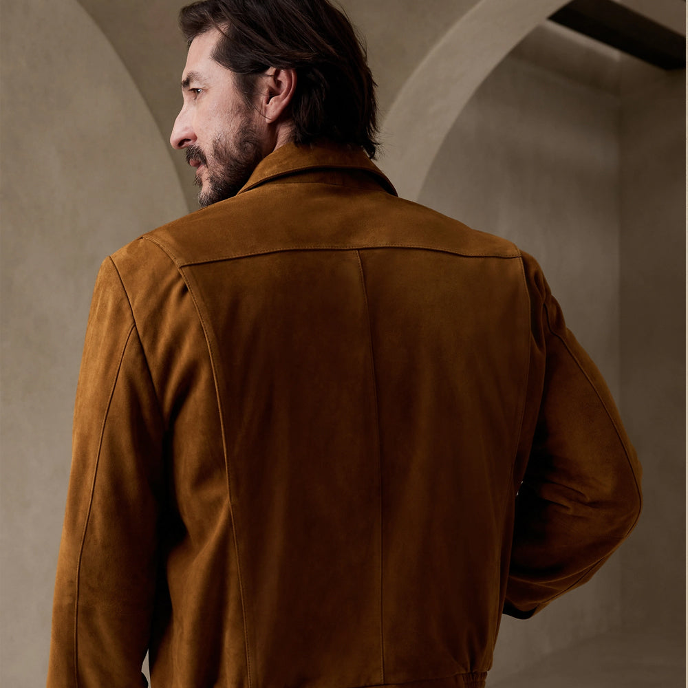 A man wearing a brown suede jacket, shown from the back in a warm, softly lit interior with arched walls.