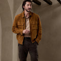 A man wearing a brown suede jacket over a beige polo shirt and dark cargo pants, standing in a rustic interior with textured walls and arches.