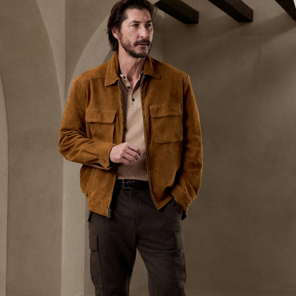 A man wearing a brown suede jacket over a beige polo shirt and dark cargo pants, standing in a rustic interior with textured walls and arches.