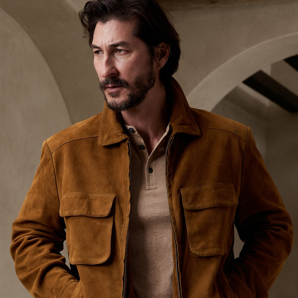 A man wearing a brown suede jacket over a beige henley shirt, standing in a softly lit architectural setting with arches.