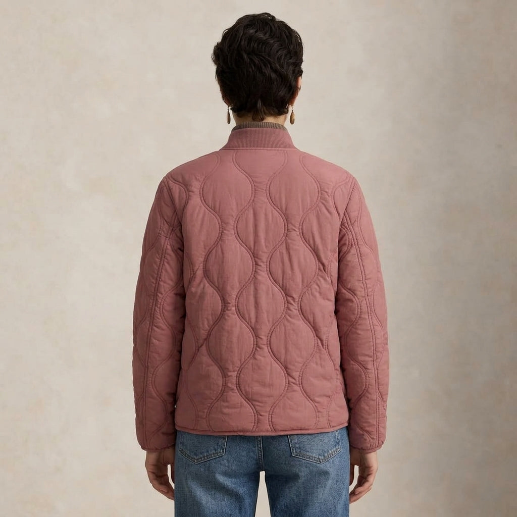 A person viewed from the back wearing a dusty rose quilted jacket and blue jeans, standing against a textured beige studio background.