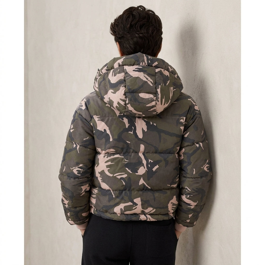 A man viewed from behind wearing a camouflage-patterned puffer jacket with a hood, paired with black pants, standing against a textured light gray wall.