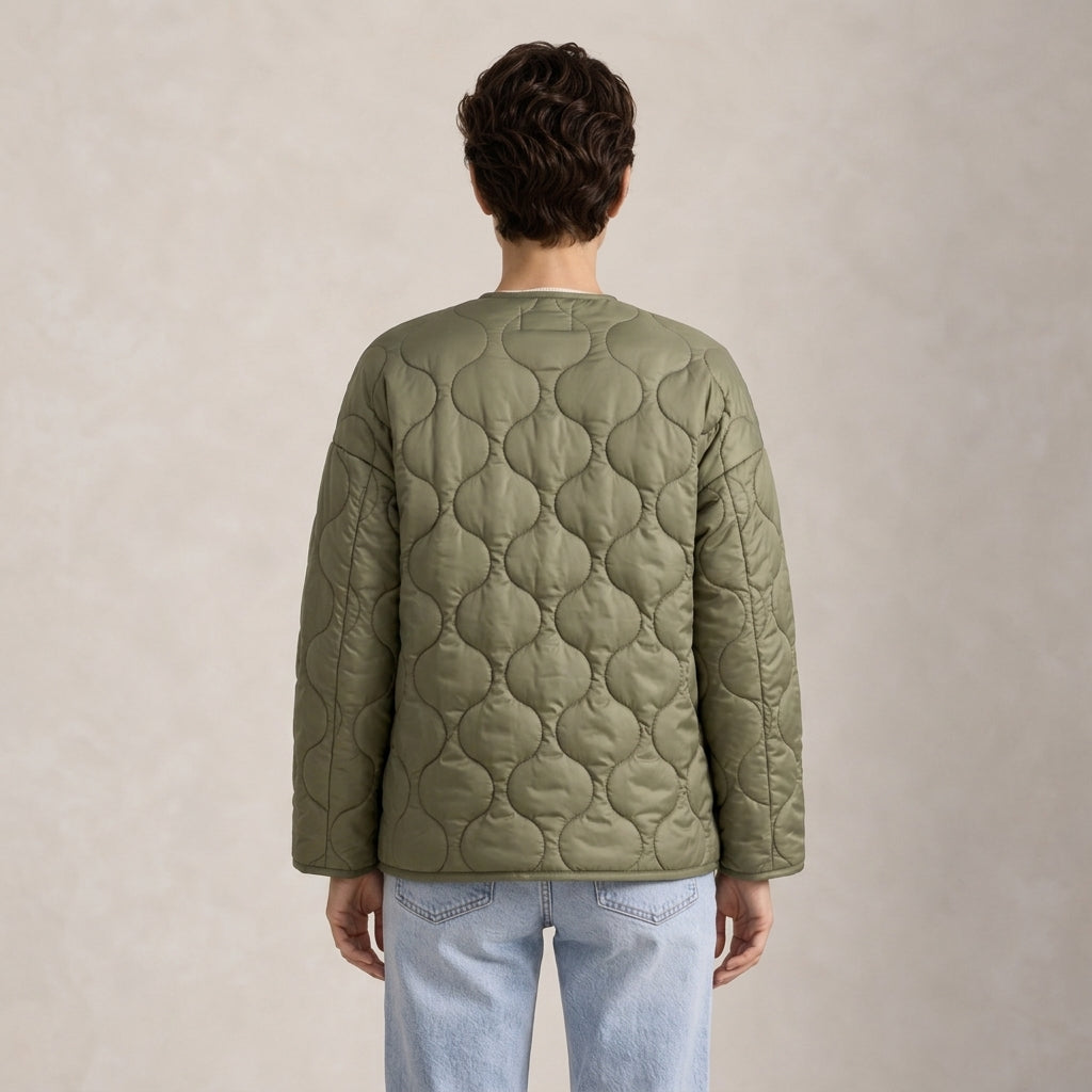 A person viewed from the back wearing an olive green quilted jacket and light blue jeans, standing against a plain textured beige studio background.