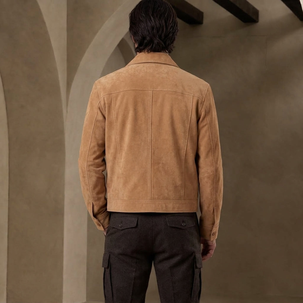 A man viewed from behind wearing a tan suede jacket and dark cargo pants, standing in a neutral interior with textured walls and arches.