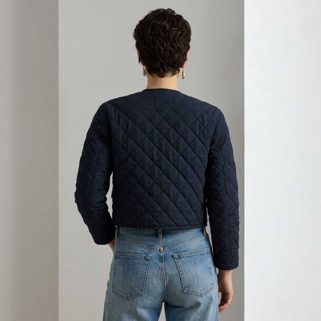 A person shown from the back wearing a navy quilted cropped jacket with long sleeves and light blue jeans, standing against a minimalist light gray wall.