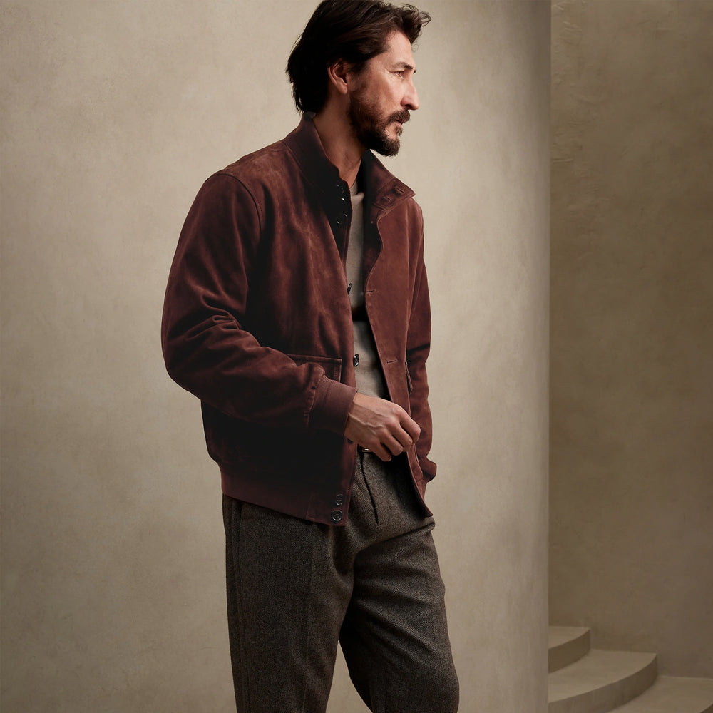 A man wearing a dark brown suede jacket over a black shirt and gray trousers, standing in a minimalist beige architectural setting.