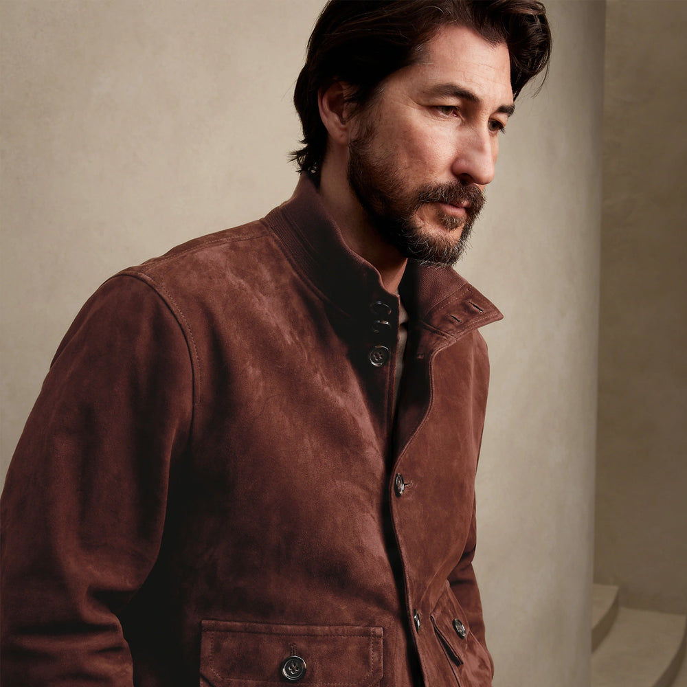 A man wearing a brown suede jacket, shown in a close-up profile portrait against a neutral beige textured wall.