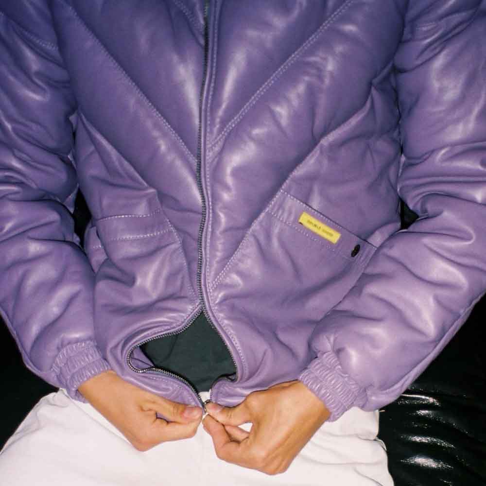 A close-up of a person wearing a shiny lilac quilted jacket, pulling the front zipper closed with both hands. The jacket has elastic cuffs, a diagonal chest pocket, and a small yellow brand tag on the pocket.