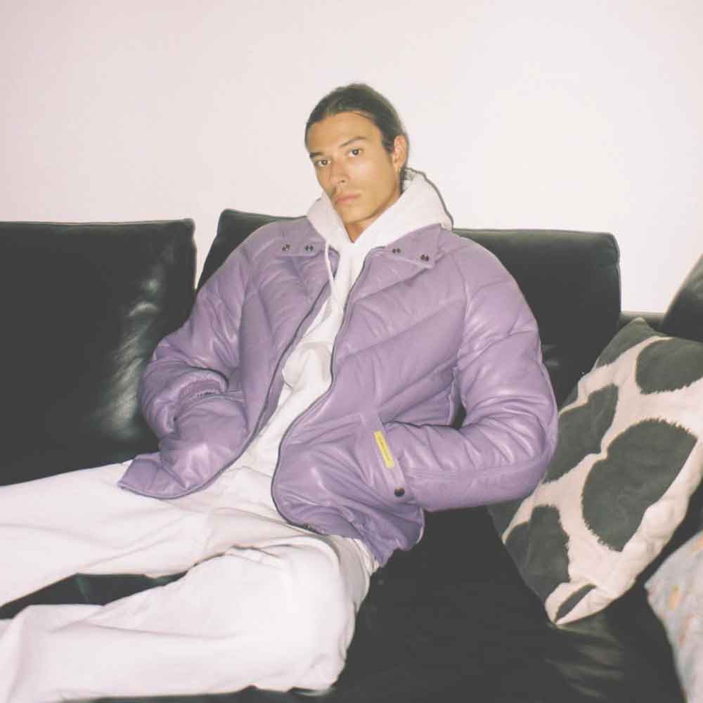A man wearing a shiny lavender v bomber jacket over a white hoodie and white sweatpants, sitting on a black leather couch with a patterned pillow.