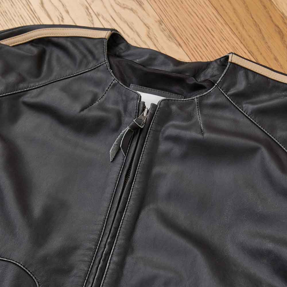 Close-up of a black leather jacket with a front zipper, laid flat on a light wood floor. The jacket has a stand collar, visible contrast stitching, and a small white tag near the collar.